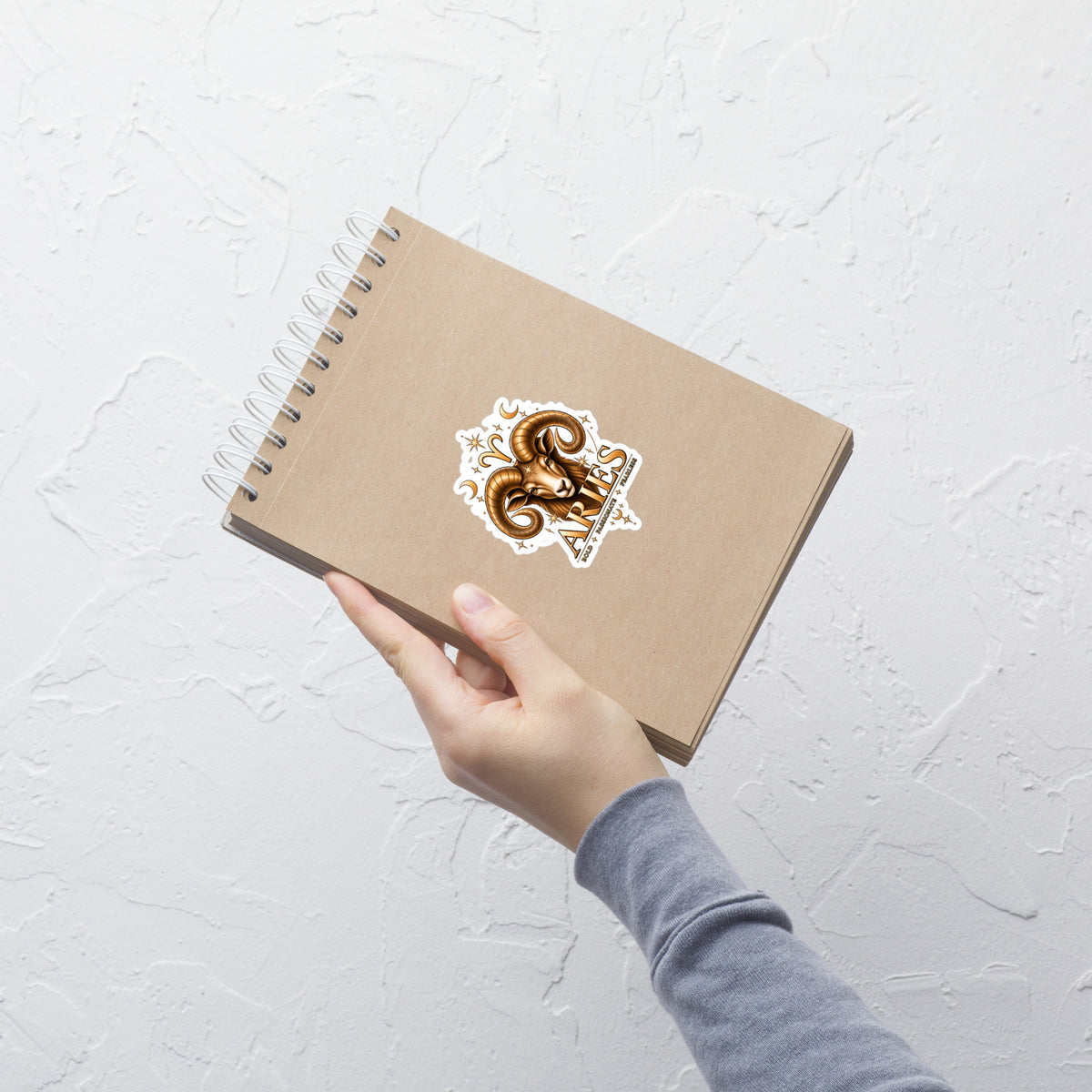 Aries zodiac sticker, 4 x 4 in kiss-cut: a golden ram with curled horns and a star on his brow in a night sky of gold stars, shown on a surface