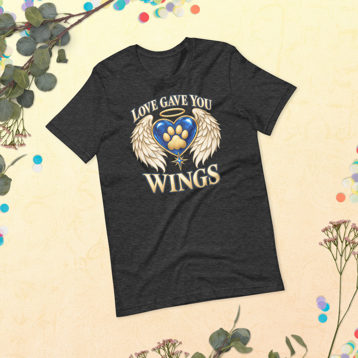 Love Gave You Wings design, a blue gem heart with a gold halo, cream angel wings and a gold cat paw print at its center, under gold lettering, on a dark grey heather unisex t-shirt, styled flat lay with eucalyptus and dried flowers on a cream backdrop