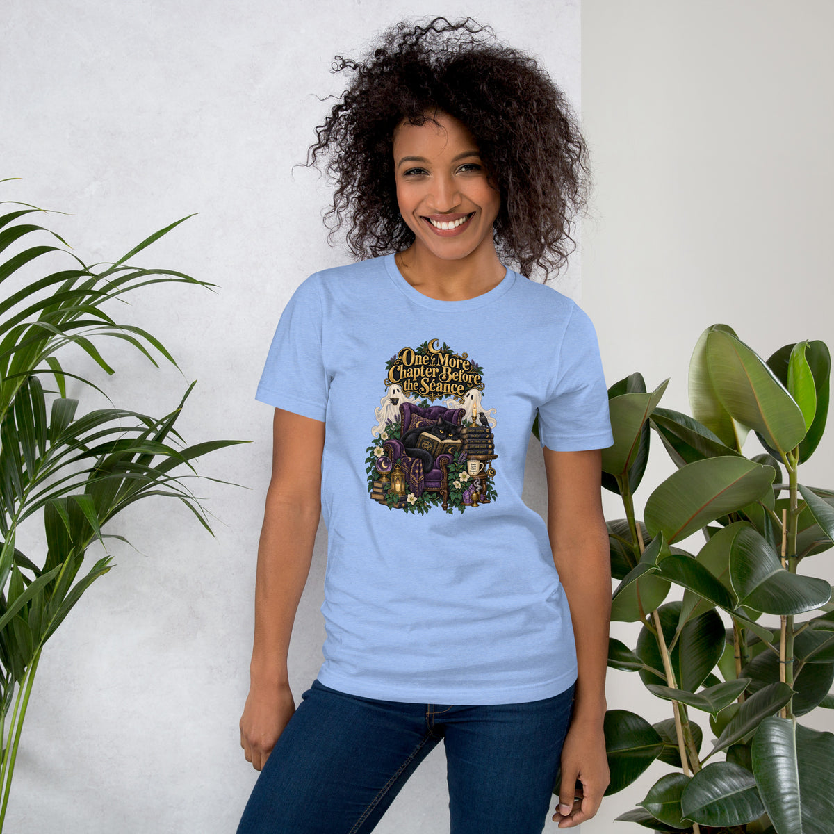 One More Chapter Before The Séance dark academia design, a black cat reading in a velvet armchair with two ghosts, on a heather carolina blue unisex t-shirt, front view