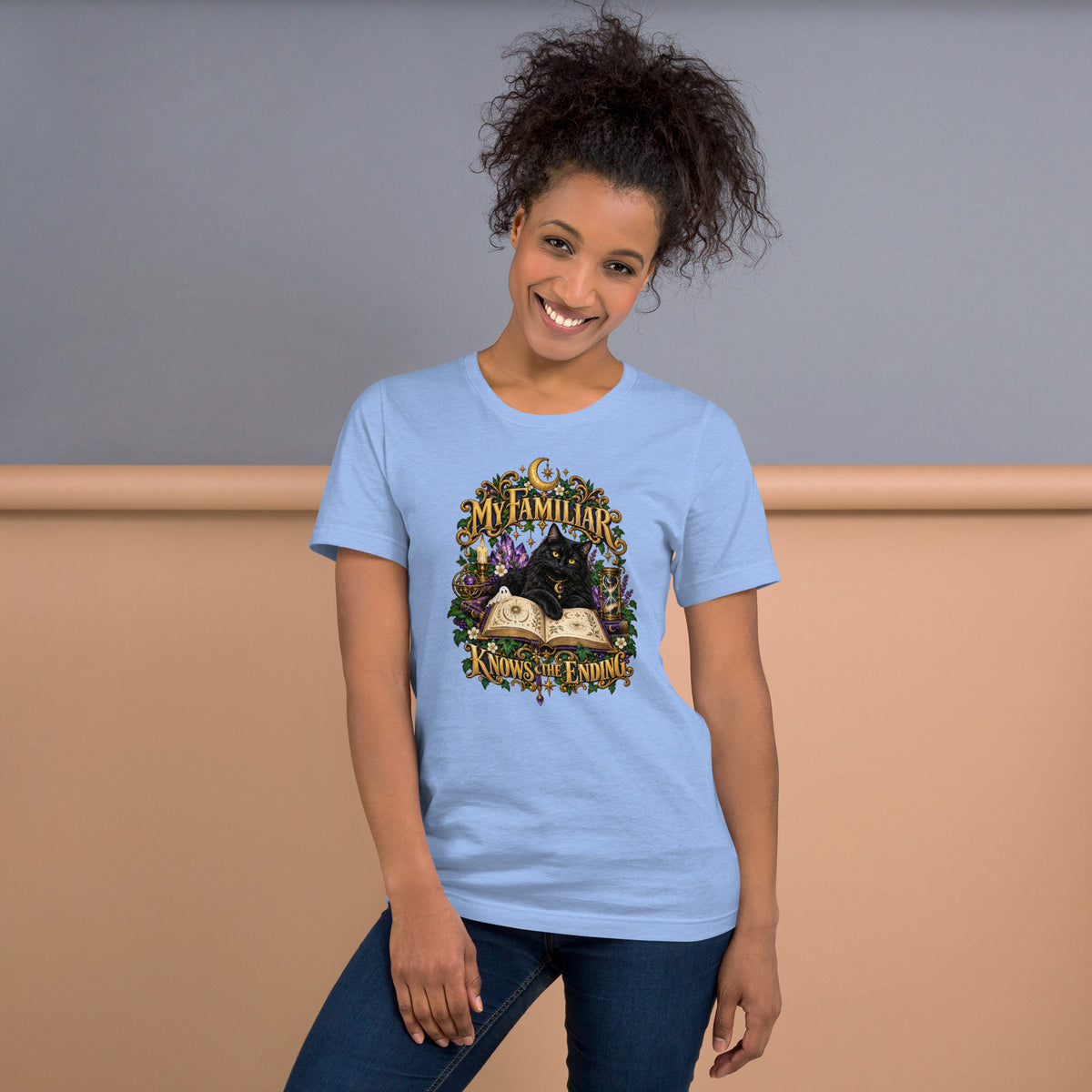 Black cat familiar on a grimoire, witchy celestial design on a heather carolina blue t-shirt, front view