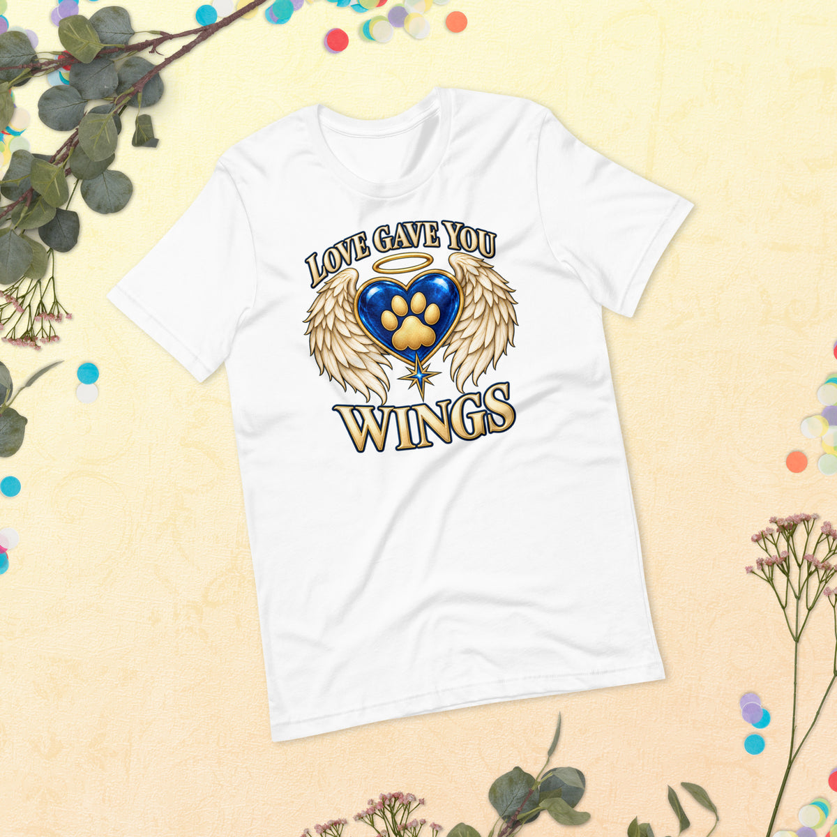 Love Gave You Wings design, a blue gem heart with a gold halo, cream angel wings and a gold cat paw print at its center, under gold lettering, on a white unisex t-shirt, styled flat lay with eucalyptus and dried flowers on a cream backdrop