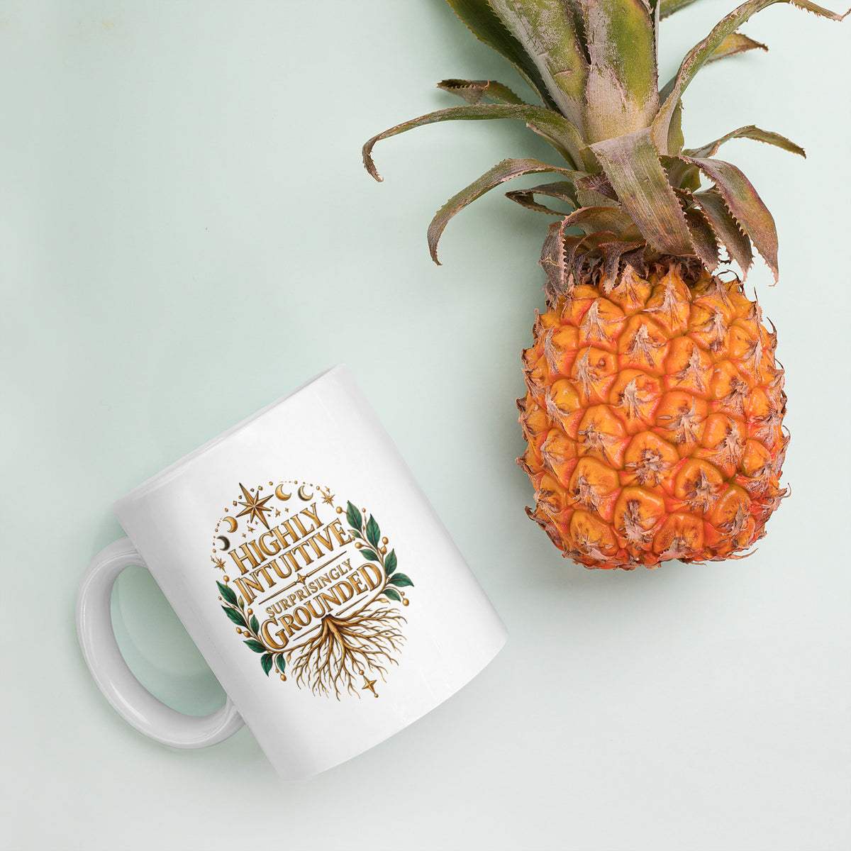 Highly Intuitive, Surprisingly Grounded design, moon phases above and roots below, on a white glossy mug, 11 oz, styled with pineapple