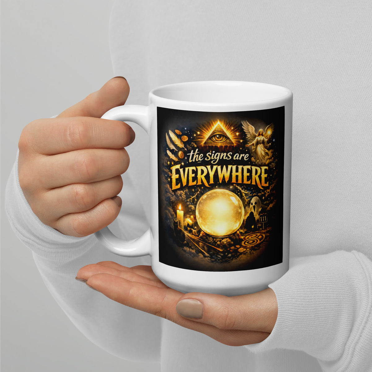 The Signs Are Everywhere design, an all-seeing eye, an angel, a glowing crystal ball, a ghost and a 111 road sign, in gold on black, on a white glossy mug, 15 oz, handle on the left